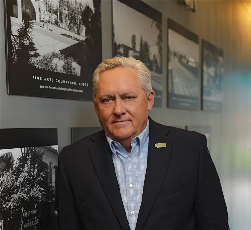 Dr. Kevin G. Walthers, superintendent/president of Allan Hancock College, stands indoors wearing a dark suit jacket over a light blue checkered shirt. He has short gray hair and a calm expression. Behind him on the wall are framed black-and-white historical photos of the college campus.